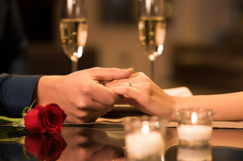 A closeup of a couple holding hands on a restaurant table with two glasses of champagne in background. There are also candles and a rose on the table.