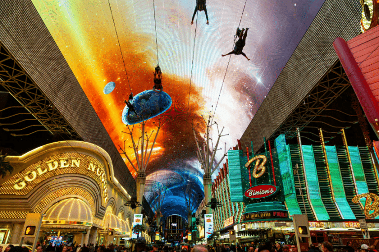 a lighted canopy photo of the Fremont Street Experience as zipliners go by. Beneath the ziplines is a large crowd standing outside the Golden Nugget which is a hotel and casino.