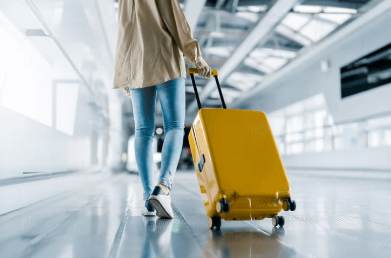 an image of the back of a woman who is wheeling a yellow suitcase behind her in an airport as she takes a vacation.